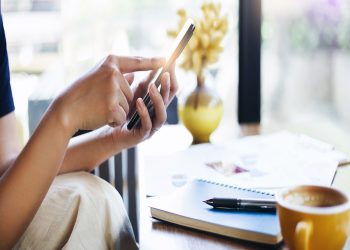 Close-up young asian woman touching on mobile screen to order food while she work at home with notebook and coffee drink on desk