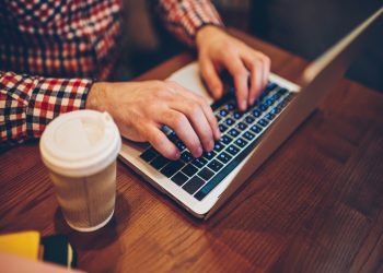 Cropped image of male hands typing information for website via keyboard on laptop computer connected to free wireless internet in coffee shop, it professional working remotely with modern netbook