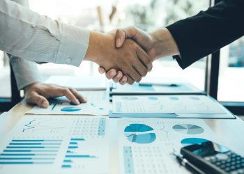 Entrepreneurs collaboration deal shaking hands in a modern office and financial paper graph on desk.