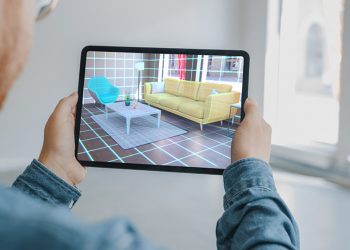 Decorating Apartment: Man Holding Digital Tablet with AR Interior Design Software Chooses 3D Furniture for Home. Man is Choosing Sofa, Table for Living Room. Over Shoulder Screen Shot with 3D Render