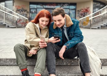 Two teenagers holding smartphones, looking at the screen, discussing something and smiling while sitting on the steps outdoors. Technology, smartphone addiction. Selective focus. Horizontal shot
