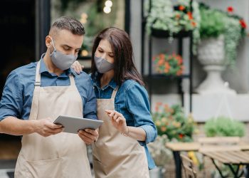 Small business and work with gadgets online during covid quarantine. Millennial guy and woman in aprons and protective masks look at tablet and take online order near flower shop outdoor, copy space