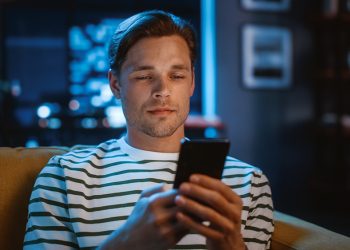 Handsome Caucasian Man Using Smartphone in Cozy Living Room at Home Sitting on a Sofa in Evening. Doing Online Shopping, Browsing the Internet and Checking Videos on Social Media, Remote Work